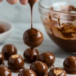 Hands dipping chilled Butterfinger balls into melted chocolate with crushed candy bars in the background