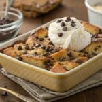 A delicious serving of chocolate chip bread pudding topped with whipped cream and extra chocolate chips, showcasing its golden, crispy edges and melted chocolate filling. Perfect for dessert lovers!